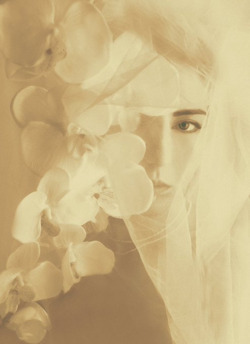 The image features a woman with long hair wearing a veil, which has white orchid petals attached to it. The background of the photo appears to be a gray color. There are no other objects or people visible in the image.