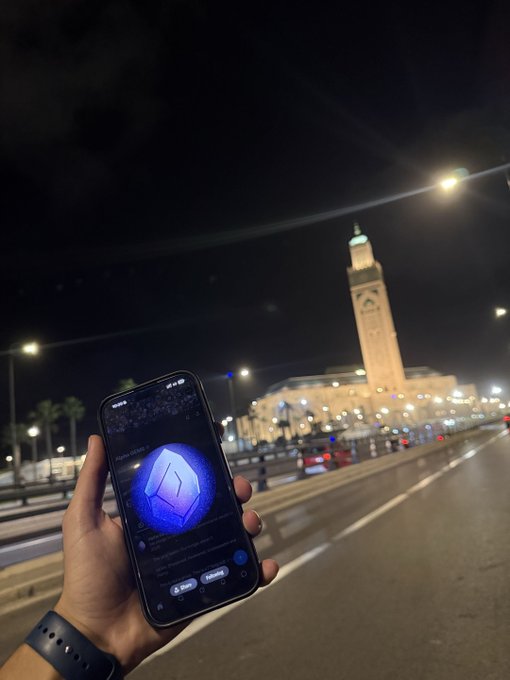 Nighttime photograph of a hand holding a smartphone displaying a purple geometric logo, with an illuminated clock tower and streetlights visible in the background.