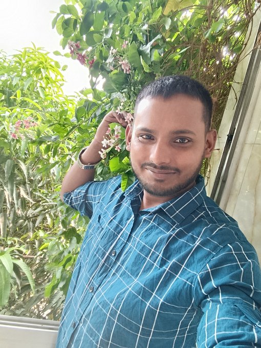 A man with a short beard wearing a teal checkered shirt takes a selfie on a balcony with green foliage in the background.