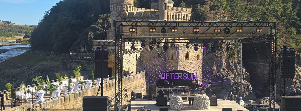 An outdoor stage is set up with lighting rigs and a banner reading '@FTERSUN' in pink neon letters, located in front of a castle with a river visible in the background.