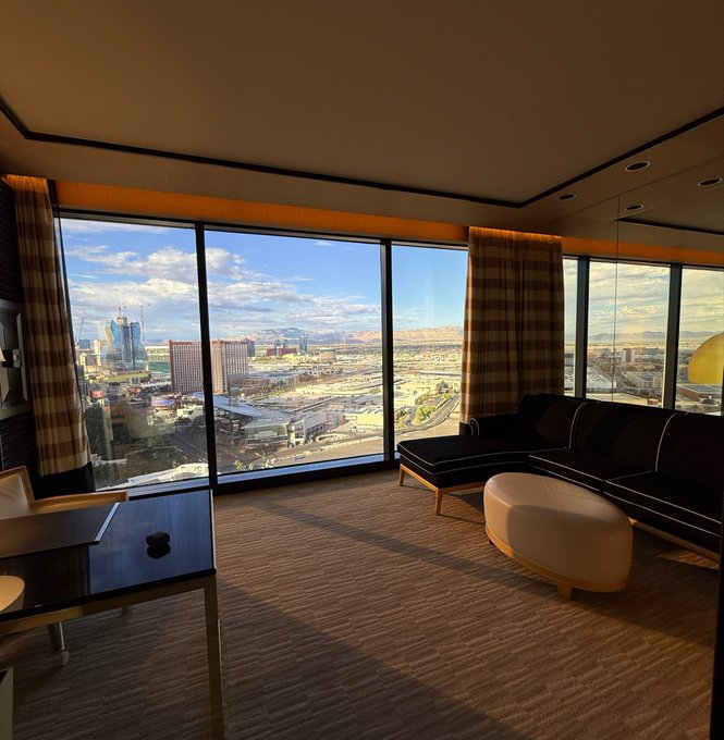 An interior view of a high-rise hotel room or suite with floor-to-ceiling windows overlooking a city skyline, featuring a couch, ottoman, and coffee table with a view of Las Vegas-style buildings in the distance.