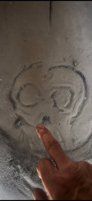 A hand points to a skull drawing etched in the sand on a beach.