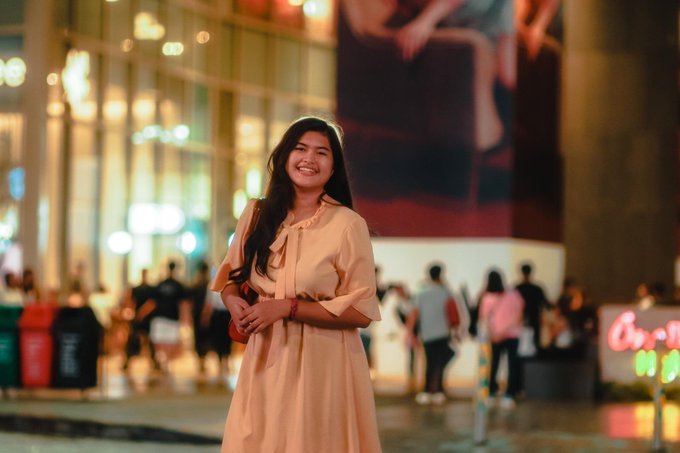 A smiling young woman with long dark hair poses in a peach-colored dress on a city street at night with bokeh lights in the background.