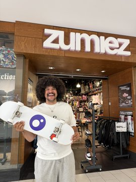 A person with an afro and a mustache holds a skateboard in front of a Zumiez store. The skateboard features a circular logo with a purple outline and a blue dot.