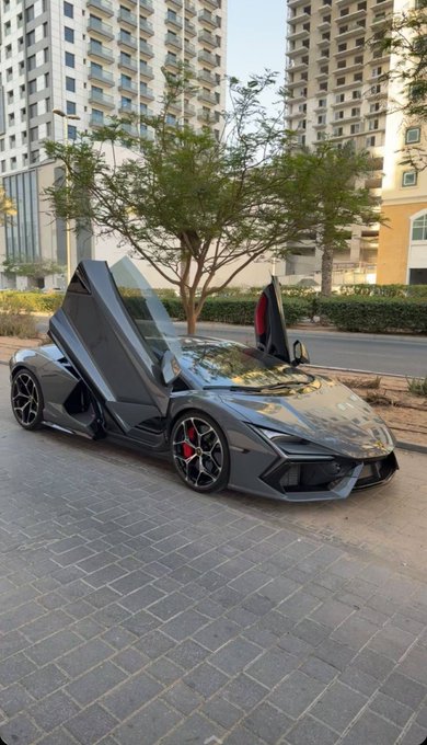 A sleek gray Lamborghini sports car is parked on a paved driveway with its signature scissor doors open upward, displayed against a backdrop of modern high-rise buildings and trees, perfectly matching the post's confident "GM to everyone who says GM" message by showcasing a luxurious supercar that demands attention.