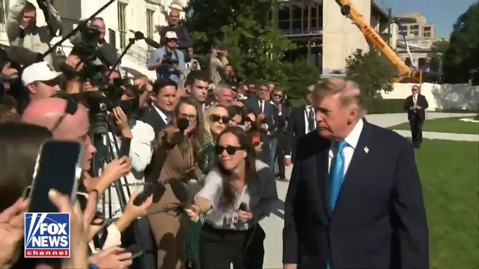 Donald Trump in a dark suit and blue tie walks past a crowd of reporters and photographers outside a building, with a "FOX NEWS channel" logo visible in the bottom left corner.