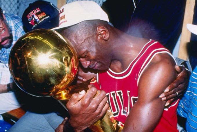 Michael Jordan, wearing his red Chicago Bulls jersey and a white championship hat, embraces the golden NBA Championship trophy while kneeling amid a locker room celebration.