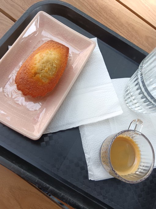 A tray holds a square ceramic plate with a small pastry or madeleine, accompanied by a glass of water and a glass cup of espresso on a paper napkin, set on a wooden table.
