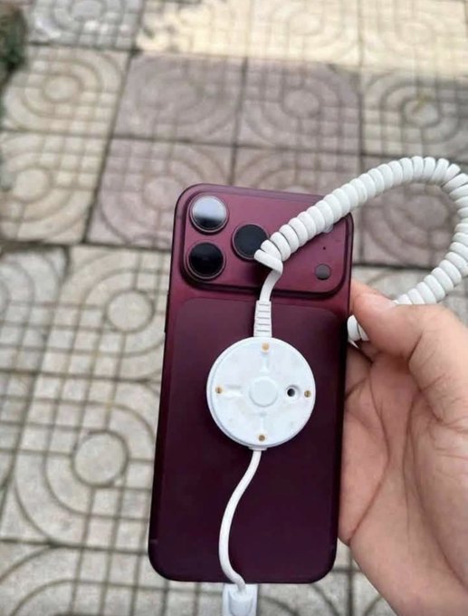 A hand holding a purple iPhone with a white circular magnetic charging attachment stuck to its back, connected to a coiled white cable, with tiled pavement in the background.