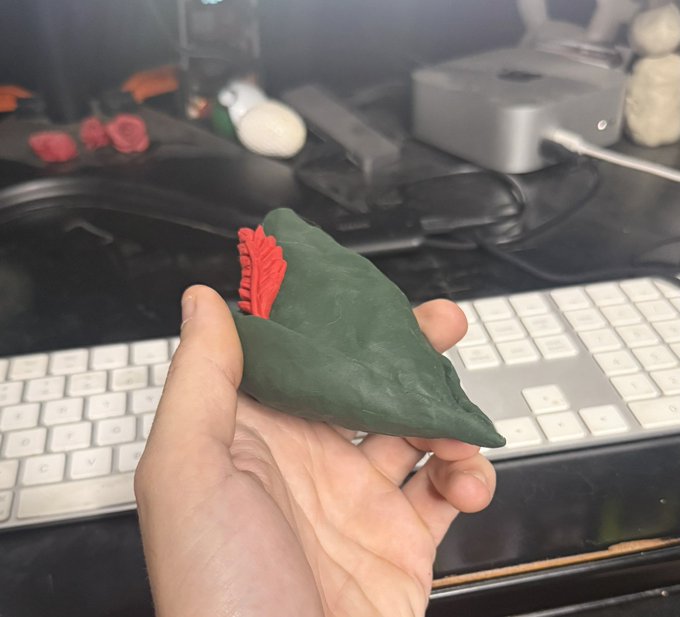 A hand holding a green leaf-shaped clay or plastic figurine with red details, held over a desk with a white Apple keyboard in the background.