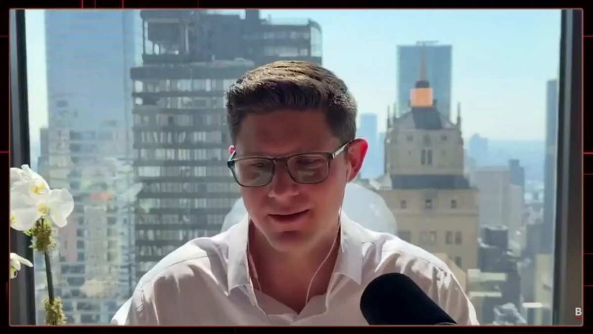The image shows an urban cityscape, featuring tall buildings at various heights against a clear blue sky. The main subject of the picture is a man wearing glasses who appears deep in thought or possibly pondering something important while sitting by his window overlooking the bustling city outside.