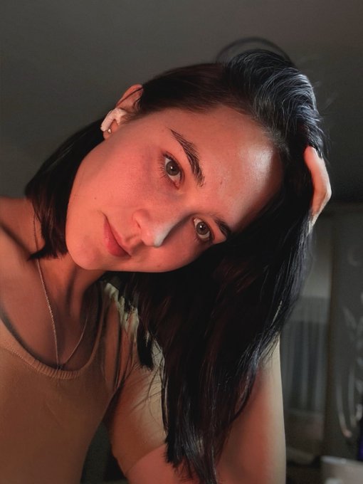 A close-up selfie of a young woman with dark hair, wearing a light-colored top and a delicate necklace, posing with her hand behind her head.