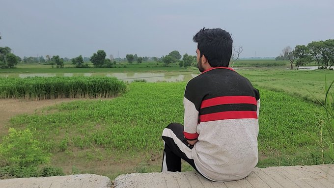 A young man in a black, red, and white jacket sits on a ledge overlooking a lush green field with water and trees in the distance.