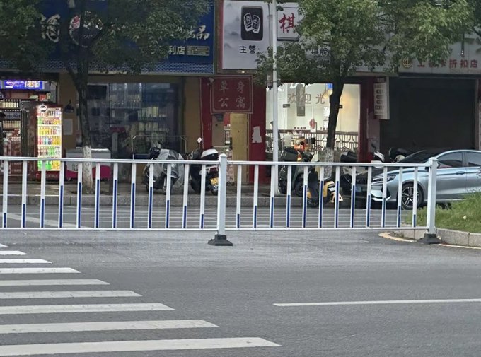 A street scene showing white and blue traffic barriers along a road with a crosswalk, lined with shops displaying Chinese characters, parked scooters, and a car.