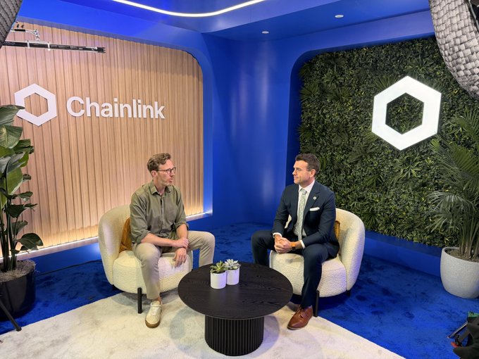 Two men sitting in armchairs having a conversation in a branded Chainlink studio with blue walls, wood paneling, and the Chainlink logo displayed prominently.