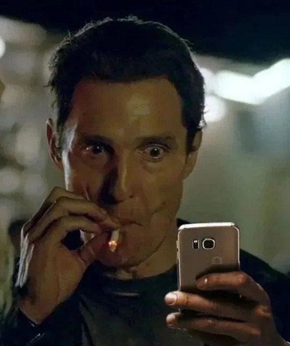 Matthew McConaughey looks at his smartphone with wide eyes while smoking a cigarette, in a scene that appears to be from a film.