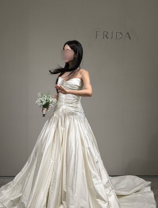 A woman models a lustrous ivory satin wedding gown with a strapless sweetheart neckline and a dramatic ruched bodice flowing into a voluminous skirt, holding a small bouquet of white baby's breath. The brand name "FRIDA" appears in dark lettering on the wall behind her, and the caption "My best mint ever." playfully refers to the pristine, untouched condition of the gown as if it were a freshly minted treasure.