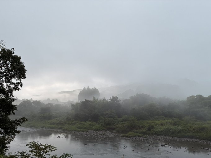 A moody landscape photograph showing a misty river flowing through a forested valley, where fog obscures distant buildings or a castle on the hillside under an overcast sky.