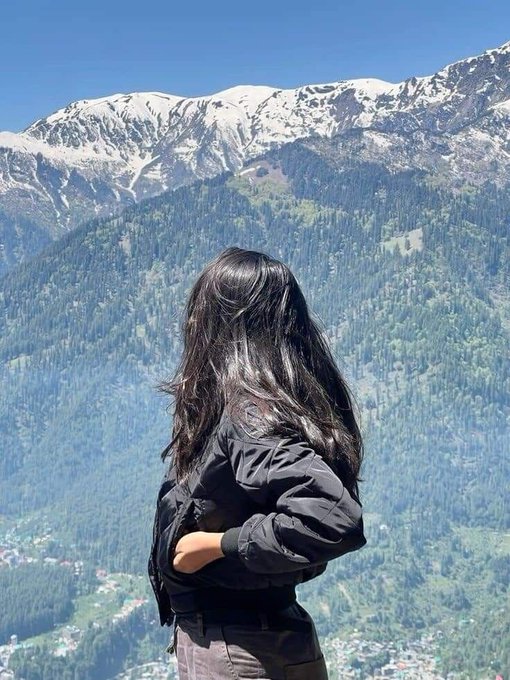 A woman with long dark hair stands with her back to the camera, looking out at a scenic view of forested mountains with snow-capped peaks.