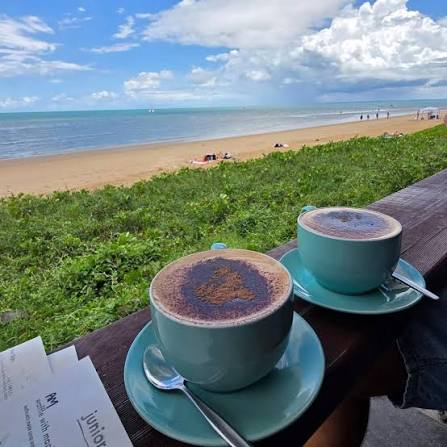 Two cappuccinos dusted with cocoa powder sit on teal saucers at a seaside café table, overlooking a sandy beach with people sunbathing and a partly cloudy blue sky. The cozy breakfast view complements the "gm" (good morning) post perfectly, suggesting a relaxing morning spent enjoying coffee by the ocean.