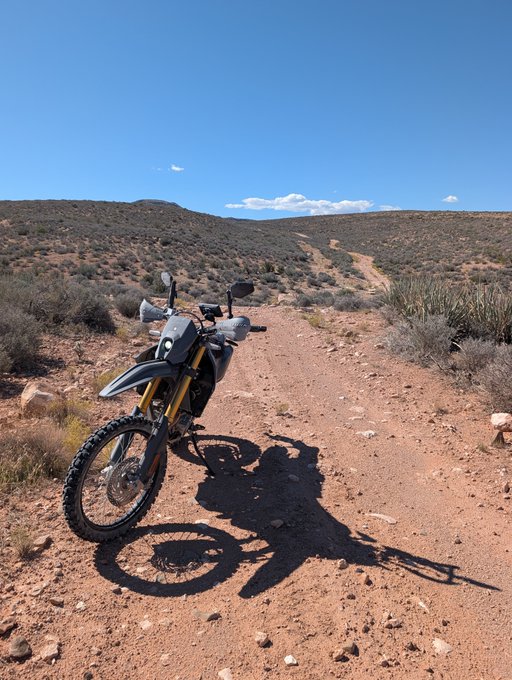 A dirt bike parked on a rocky desert trail under a clear blue sky, with the rider's shadow cast on the reddish-brown dirt ground alongside scrubby vegetation.