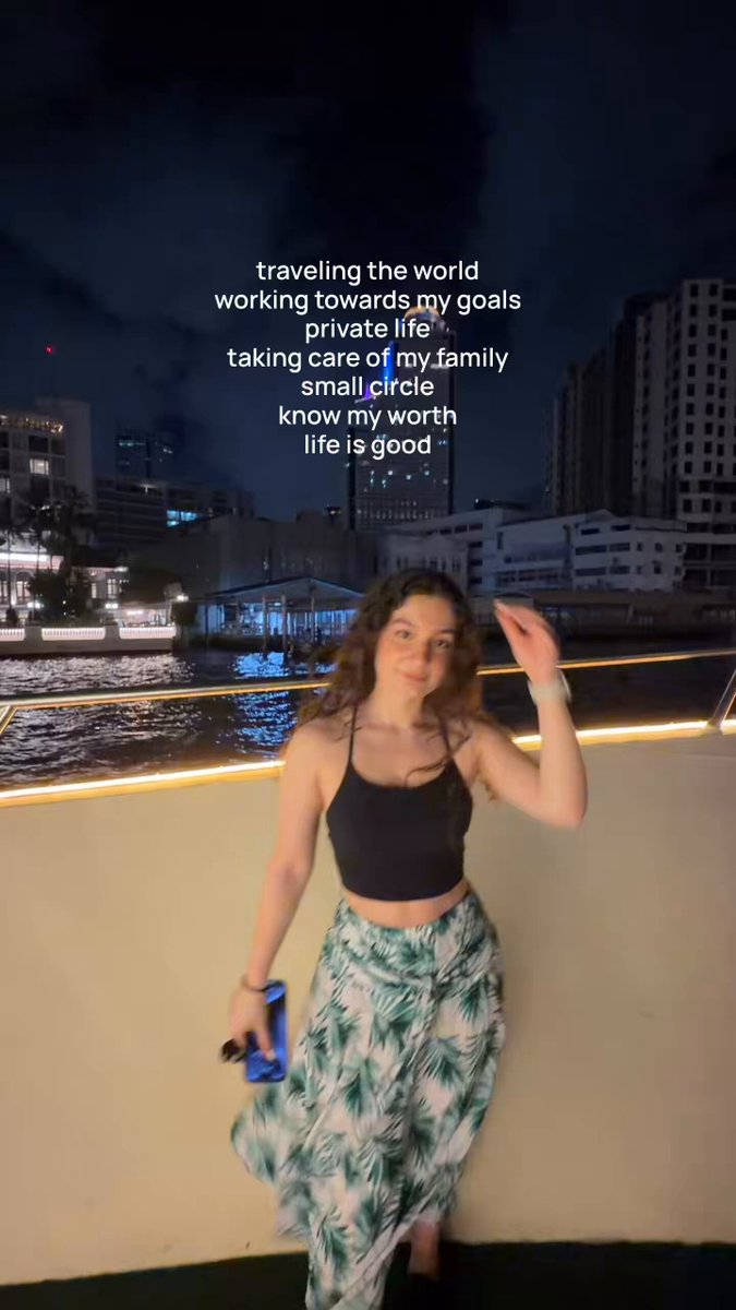 The image features a woman standing next to a building, wearing a black tank top with green leaves printed on it. She has her arm raised above her head as if posing for a photo or video. The background of the image shows a city skyline at night, which adds an urban atmosphere to the scene. There is also text in the image that reads "traveling the world towards my goals", suggesting that she might be on a trip or vacation.