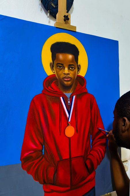 An artist paints a portrait of a young Black man with a halo, wearing a red hoodie and a bronze medal on a ribbon, set against a bright blue background.