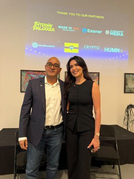 A man and woman pose together at an event with a banner thanking partners including 'Bitcoin Paloma', 'Made in USA', 'Easner', 'Mebixa', 'Blocknation', 'Crypto', and 'Humann'.