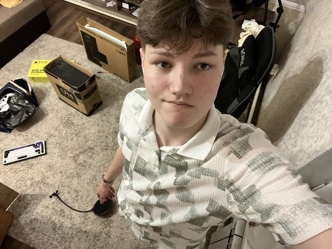 A young person wearing a green and white patterned polo shirt takes a selfie in a living room, with several shoe boxes (including Hoka and Reebok) and a sneaker scattered on the carpet behind them.