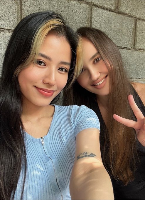 A selfie of two smiling young women, one with blonde highlights in a light blue ribbed top and the other with long brown hair in a black top making a peace sign.