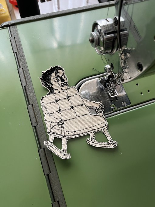 An embroidered fabric patch depicting a man transformed into a tufted armchair is being sewn onto a green fabric surface using a sewing machine. The playful craft visually matches the caption's whimsical concept of "Eloy" being turned into a chair as a result of saying he's tired.