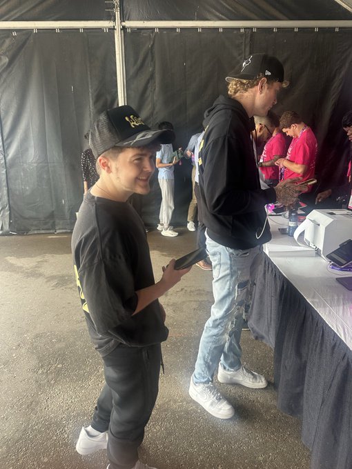 Photo of two young men wearing black caps and hoodies, standing under a black tent while looking at their phones near a table with merchandise.