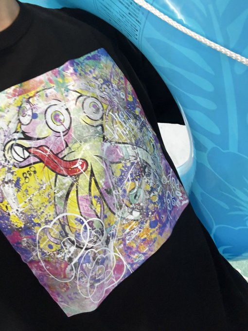 A person wearing a black t-shirt featuring a colorful, abstract print with a cartoon face sticking its tongue out, set against a turquoise inflatable pool.
