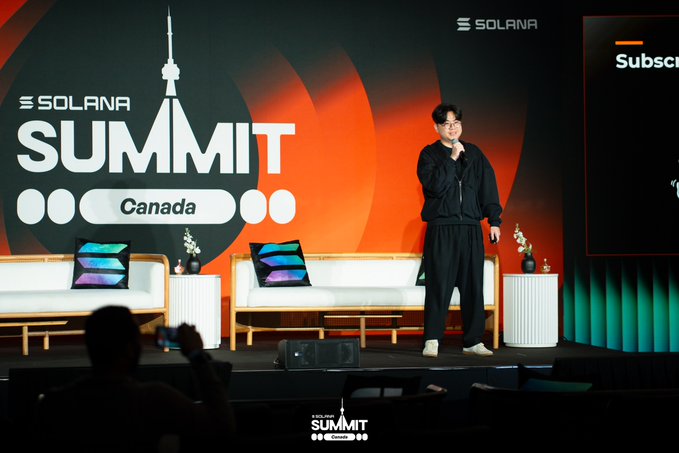A man stands on stage speaking at the 'SOLANA SUMMIT Canada' event, with the Solana logo prominently displayed on the backdrop alongside a stylized city tower graphic.