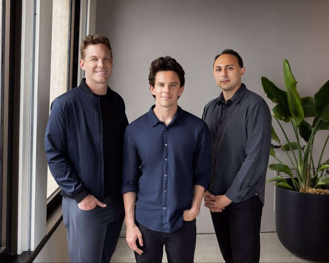 Three men stand together in a modern, plant-accented office space—the central figure in a navy button-up shirt flanked by a taller man in a bomber jacket and another in a dark button-down, posing for what appears to be a professional team photo. The image ties to the post's narrative of an industry connection, with the central man likely being the "Rajiv" referenced as having advanced to GP (General Partner) since their casual Blue Bottle coffee meeting years ago.