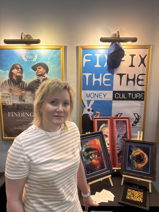 In the image, a woman wearing a white shirt stands next to several framed posters of movies or other artwork. She appears to be posing for a photo with these posters behind her. The poster she is standing in front of has the words "Fix It" written on it. There are also two small paintings displayed nearby, one of which features a cat and the other an abstract design. A book can be seen resting on a surface next to the woman as well.