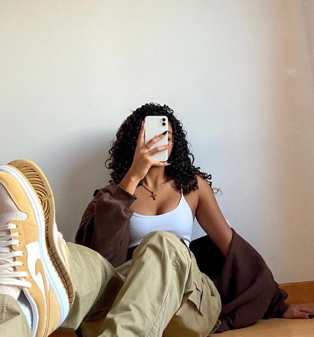 The image features a woman sitting cross-legged on the floor while holding her phone up to take a selfie. She has curly hair, which she is capturing in the photo. The background of the image shows a white wall with a pair of shoes placed against it. There are no other people or objects visible in the scene.