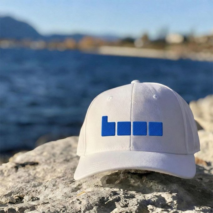 A white baseball cap with a blue 'bom' logo resting on a rock by a lake or coastline.