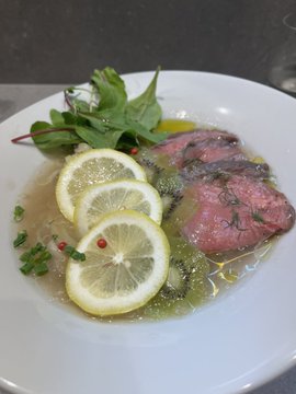 A plated dish featuring slices of rare roast beef covered in a green herb sauce, garnished with lemon slices, pink peppercorns, and fresh greens.