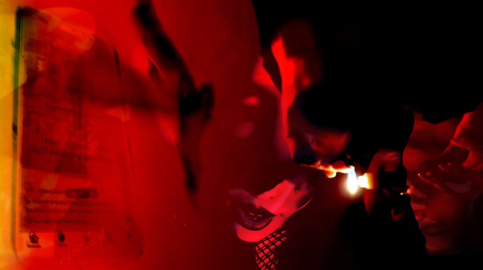 A dimly lit, red-tinted scene of a person smoking, with their reflection visible in a mirror or glass surface.