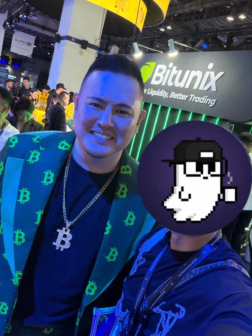 A man wearing a Bitcoin-patterned scarf and a Bitcoin pendant necklace poses in front of a 'Bitunix' booth at a crypto conference, with another person's face obscured by a pixel art avatar.