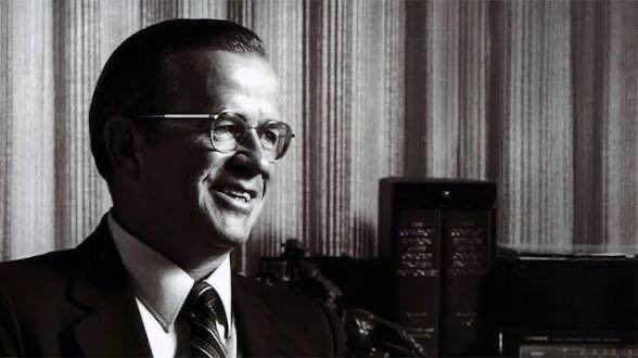 A black and white vintage photograph of a smiling man wearing glasses, a suit, and tie, appearing to be in an office setting.