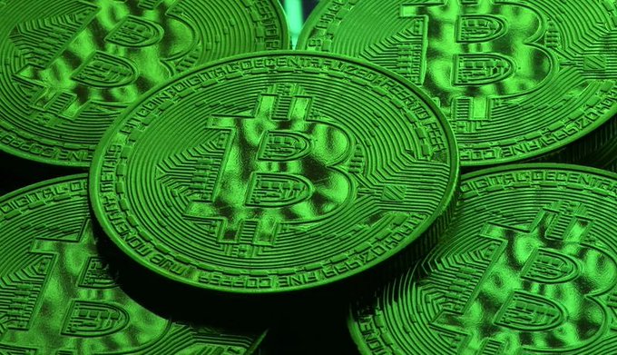 A close-up photograph of a pile of physical Bitcoin tokens illuminated with green lighting.