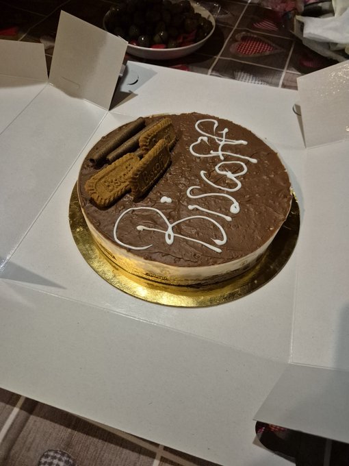 A round chocolate cake in a white box, decorated with white icing writing and topped with chocolate-covered biscuits and wafer rolls, with a bowl of olives visible in the background.