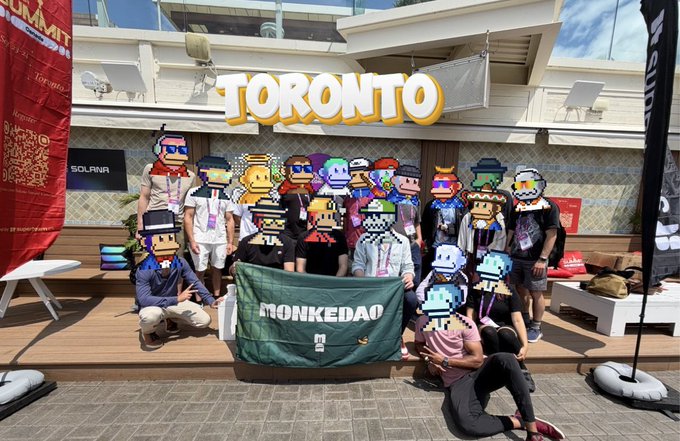 A group of attendees pose together at the Futurist Conference in Toronto, holding a green "MONKEDAO" banner, with their faces covered by pixel-art avatars in front of a large illuminated "TORONTO" sign, capturing the community spirit of the MonkeDAO meetup.