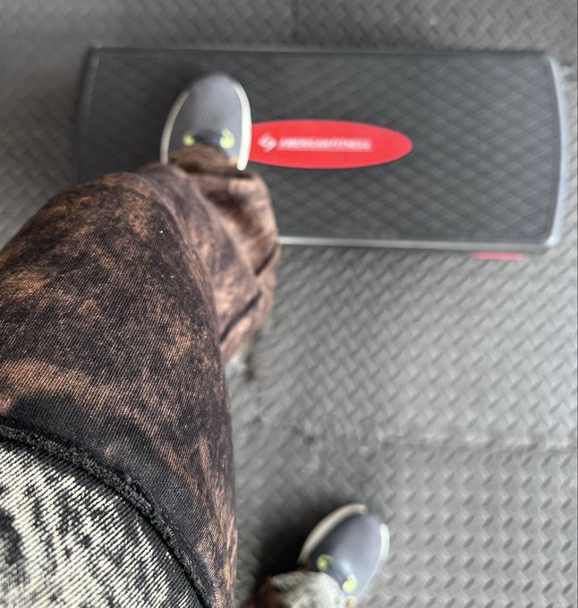 A person's leg in patterned brown leggings stepping onto a black step platform with a red 'LOADHOUSEFITNESS' label, wearing gray Crocs on a rubber gym floor.
