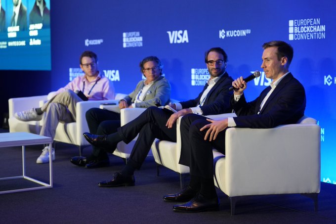 A panel discussion at the European Blockchain Convention featuring four men seated on white chairs on stage with Visa and KuCoin branding visible in the background.