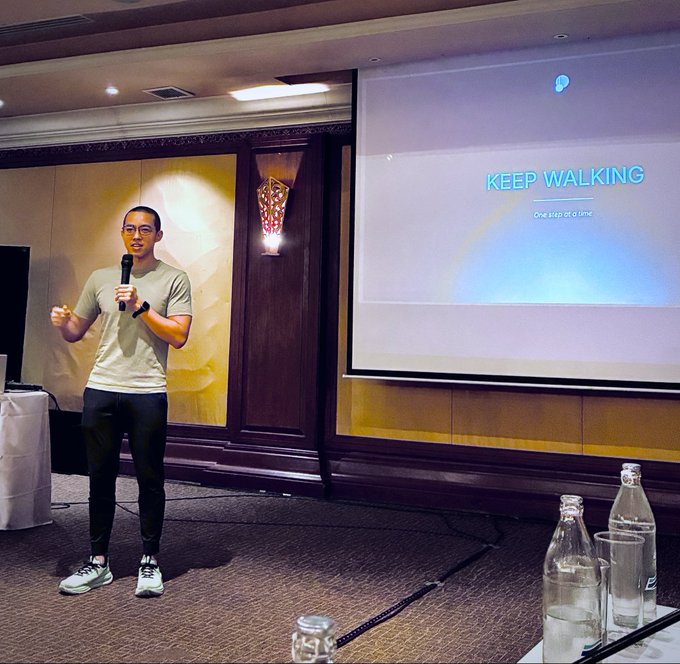 A man in casual attire stands on stage holding a microphone, presenting in front of a large screen displaying the motivational phrase "KEEP WALKING - One step at a time," aligning with the post's theme of Pendle's ongoing journey and dedication to shipping through any market conditions.