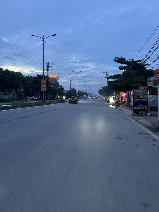 A quiet road at dawn in a small town, with a glowing red sign reading "Bến Xe" and a yellow "PC GAMING - LAPTOP - CAMERA" billboard on the right, captures the stillness of early morning before activity begins. The empty street mirrors the post's sense of waiting and being on pause, as if the town itself is holding its breath for the day to start.