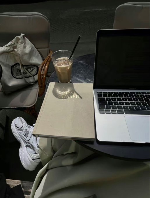 An overhead view of a coffee shop table with an iced coffee, a MacBook laptop, a 'CHASER CHICAGO' branded menu or book, a tote bag, and a person's foot in white New Balance sneakers.